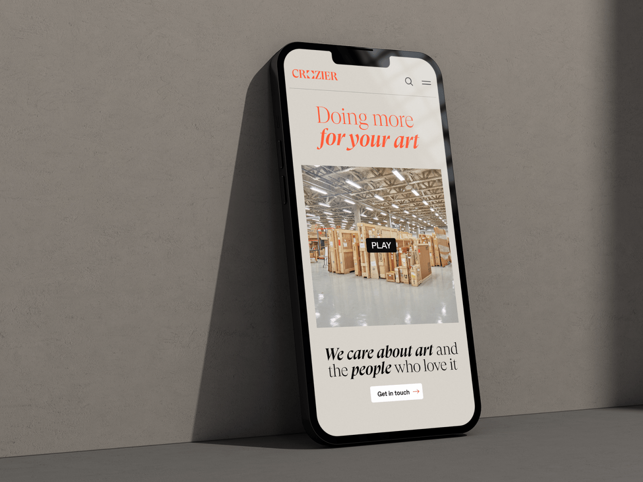 An iphone leaning against a grayish wall. On the screen is the Crozier homepage, featuring orange and black text that reads: &quot;Doing more for your art&quot; and &quot;We care about art and the people who love it.&quot;