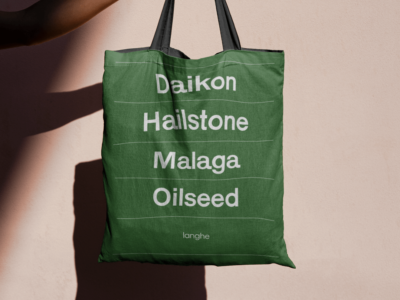 A forest green totebag being held against a tan wall. White text on the bag reads &quot;Daikon, Hailstone, Malaga, Oilseed&quot; and &quot;Langhe.&quot;
